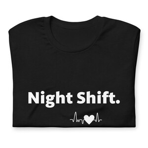 May include: Black t-shirt featuring the white text "Night Shift." and a white heartbeat graphic with a heart symbol. The t-shirt is folded neatly. A casual garment.