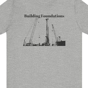 以下が含まれることがあります： 建設クレーンと機械の黒いグラフィックが特徴のヘザーグレーのTシャツ。「Building Foundations」の文字がグラフィックの上に印刷されています。このシャツはクルーネックで、柔らかい素材で作られています。