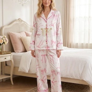 May include: White pajama set with a pink bow pattern. The long-sleeved top and pants have pink trim. The pajamas are made of a soft, silky material. The model is standing in a bedroom.