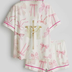 May include: White pajama set with pink bows and trim. The short-sleeved top and shorts feature a repeating pattern of pink bows and polka dots. A cross with a draped cloth is centered on the top.