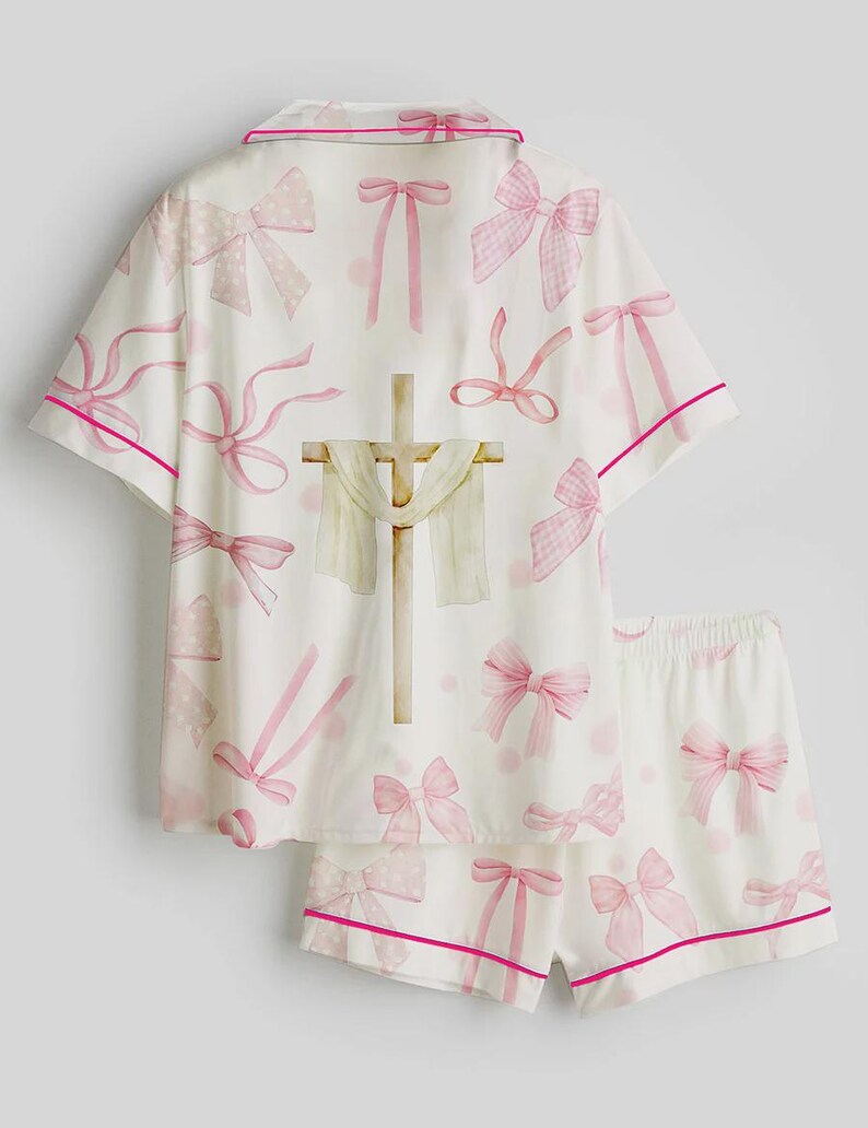 May include: White pajama set with pink bows and trim. The short-sleeved top and shorts feature a repeating pattern of pink bows and polka dots. A cross with a draped cloth is centered on the top.