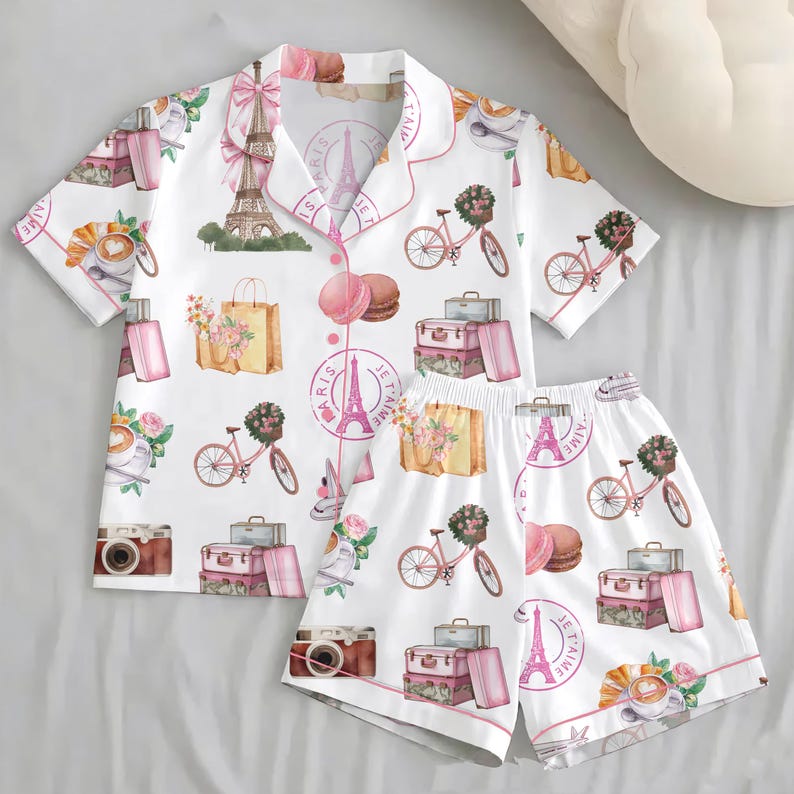 Puede incluir: Conjunto de pijama blanco con tem&aacute;tica parisina. El conjunto incluye una camisa de manga corta con botones y pantalones cortos, ambos adornados con ilustraciones rosas y beige de la Torre Eiffel, bicicletas y equipaje.