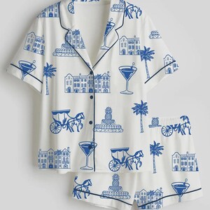K&ouml;nnte beinhalten: Wei&szlig;es Pyjama-Set mit blauem Aufdruck von Geb&auml;uden, Palmen, Cocktails und Pferdekutschen. Das kurz&auml;rmelige Oberteil und die Shorts haben marineblaue Paspeln und Kn&ouml;pfe. Ein lustiges Schlafanzug-Set zum Thema Urlaub.