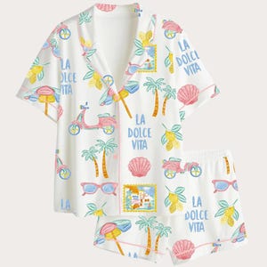 May include: White pajama set with a tropical print featuring pink scooters, palm trees, lemons, seashells, and sunglasses. The set includes a short-sleeved button-up shirt and matching shorts. Text on the set reads "LA DOLCE VITA".
