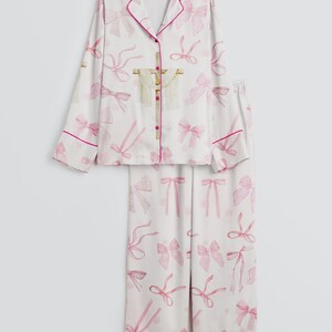May include: White pajama set with a pink bow pattern. The long-sleeved top has a notched collar and pink trim. The matching pants have a straight leg and pink trim. The set is made of a soft, lightweight fabric.
