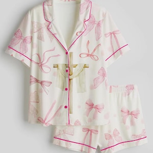 May include: White pajama set with pink bow pattern. The short-sleeved top and shorts are trimmed with pink piping. The top features a button-down front and a decorative cross design.