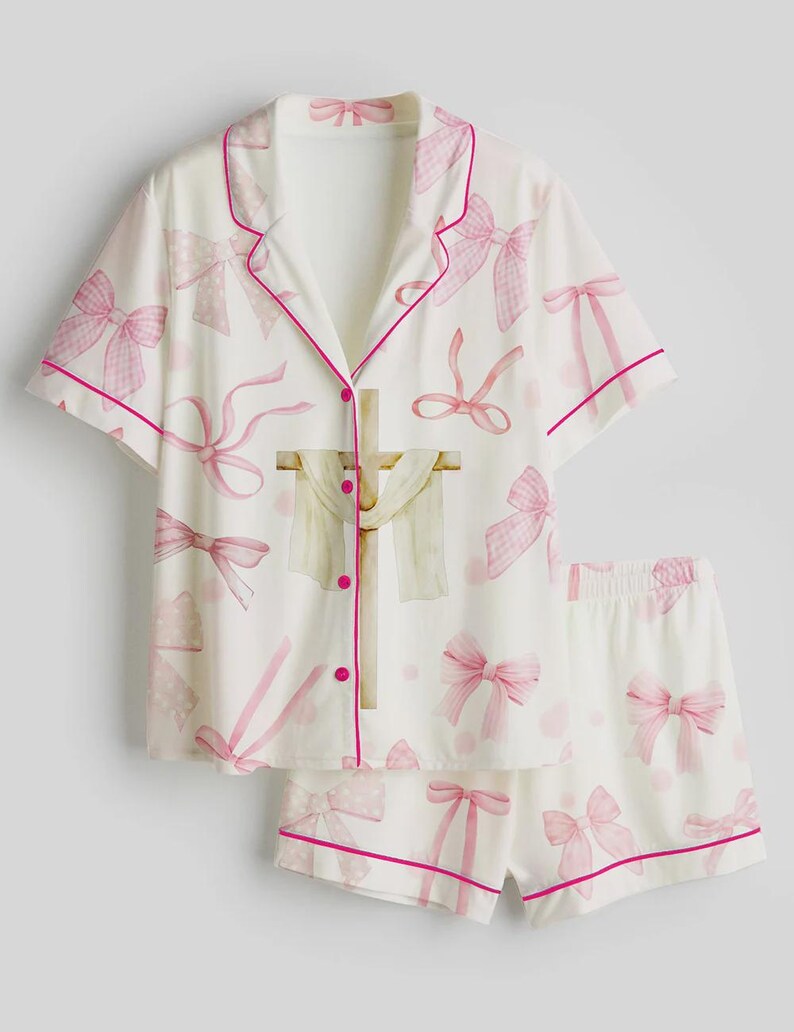 May include: White pajama set with pink bow pattern. The short-sleeved top and shorts are trimmed with pink piping. The top features a button-down front and a decorative cross design.