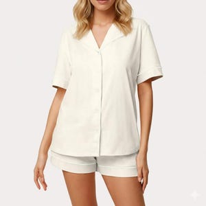 May include: Cream-colored pajama set featuring a short-sleeved button-down shirt and matching shorts. The set has white piping along the edges of the collar, sleeves, and shorts. The fabric appears to be a soft, lightweight material.