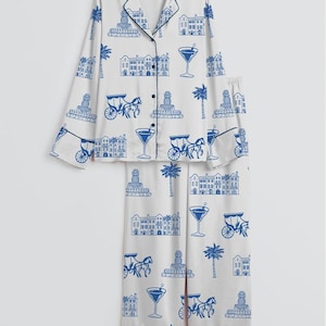 K&ouml;nnte beinhalten: Wei&szlig;es Pyjama-Set mit blauem Aufdruck, der Geb&auml;ude, Kutschen, Palmen, Ananas und Martinigl&auml;ser zeigt. Das lang&auml;rmlige Oberteil hat schwarze Paspeln und Kn&ouml;pfe, und die Hose hat einen passenden Aufdruck.