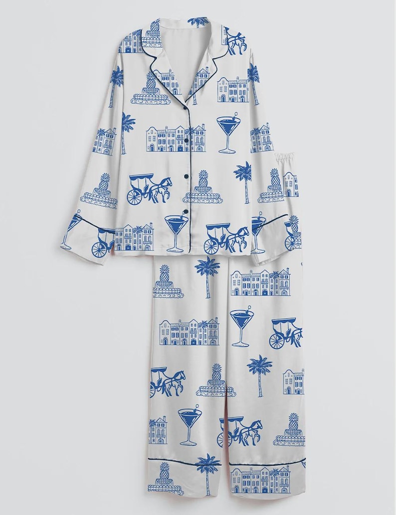 K&ouml;nnte beinhalten: Wei&szlig;es Pyjama-Set mit blauem Aufdruck, der Geb&auml;ude, Kutschen, Palmen, Ananas und Martinigl&auml;ser zeigt. Das lang&auml;rmlige Oberteil hat schwarze Paspeln und Kn&ouml;pfe, und die Hose hat einen passenden Aufdruck.