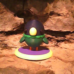 May include: A small, rounded bird figurine featuring a dark blue head, a yellow beak, and a green body with small wings. The bird stands on a circular base with a purple center and a yellow outer rim, positioned on a textured, rocky surface.