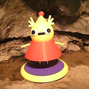 May include: A whimsical, yellow figurine with a conical head, black eyes, and a small mouth. It has a red, flared skirt and stands on a yellow, purple, and yellow circular base. The figurine has small arms and a red ball on top of its head.