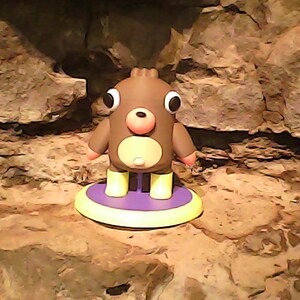 May include: A brown figurine with large white eyes, a pink nose, and a light tan belly. The character wears yellow boots and stands on a circular purple base with a yellow rim. The figure has a smooth, matte finish and a rounded, compact shape.