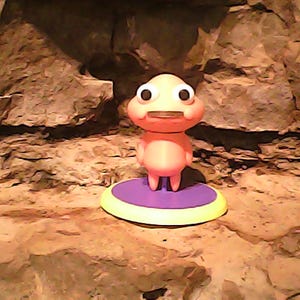 May include: A small, peach-colored figurine with a rounded head, large white eyes with black pupils, and a flat mouth. The character stands on a circular base featuring a purple center and a yellow outer rim, creating a distinct contrast in color.