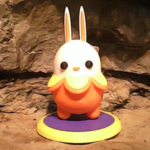 May include: A whimsical, cartoon-like rabbit figurine with a white head and long ears, accented with orange. The body is orange with a yellow and orange collar. It stands on a purple and yellow circular base. The background is a textured, rocky surface.
