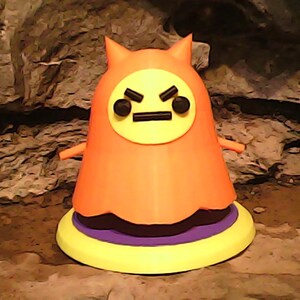 May include: A 3D-printed orange figure with a yellow face featuring black eyes and a frown. The figure has small arms and is atop a yellow, purple, and black base. The background is a textured, brown surface.