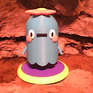 May include: A whimsical, light blue figurine with a round body, small arms, and large, cartoonish eyes. It has a red and yellow hat and sits on a purple and yellow base. The background is a textured, reddish-brown surface.