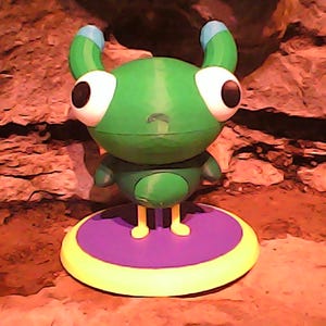 May include: A green alien-like figurine with large white eyes and blue-tipped antennae. It stands on a purple and yellow circular base. The background is a textured, reddish-brown surface, creating a whimsical, otherworldly aesthetic.