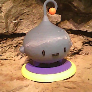 May include: A whimsical, cartoon-like figure with a bulbous, blue-gray body, small arms, and a smiling face. It has a curved antenna with an orange ball at the end. The figure rests on a circular base with yellow, purple, and yellow rings.