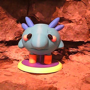 May include: A whimsical, light blue creature figurine with large, round eyes and orange legs. The figurine has blue antennae and is standing on a yellow and purple circular base. The background is a textured, reddish-brown surface.