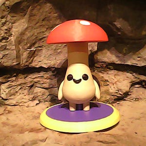 May include: A whimsical mushroom-shaped figurine with a red cap and white spots. The figurine has a cream-colored body, a smiling face, and small arms. It stands on a purple and yellow circular base. The background is a textured, rocky surface.