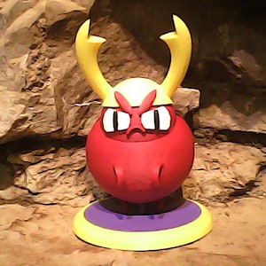 May include: A red, cartoonish figure with yellow horns and a white helmet. The figure has an angry expression with white eyes and a red mouth. It sits on a yellow, purple, and yellow circular base. The background is a rocky cave.