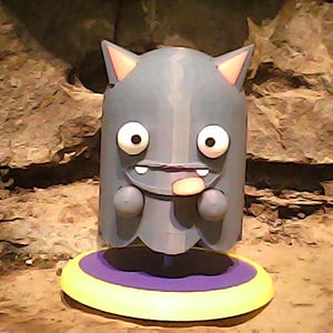 May include: A gray cartoon character figurine with large white eyes, small arms, and a protruding tongue. The figurine has pointed ears with pink accents and is standing on a yellow and purple circular base. The background is a textured brown.