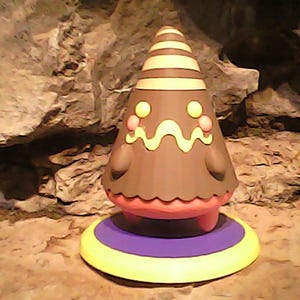 May include: A brown, cone-shaped figure with a striped top and a wavy yellow design. The figure has pink cheeks and a pink and yellow base. The background is a rocky surface.