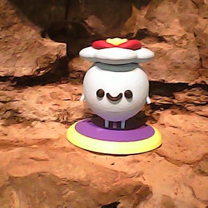May include: A spherical white figurine with a smiling face, featuring two small black eyes and a curved mouth. The figure is topped with a white cloud-like shape adorned with a red and yellow accent, and it stands on a purple and yellow circular base.