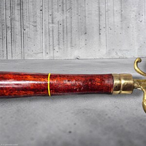 May include: A tool handle crafted from dark reddish-brown wood with a visible grain pattern, featuring a thin yellow band encircling the center and a polished brass ferrule connecting to a metallic component on the right side of the frame.