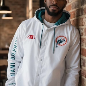 May include: White hooded jacket with a teal interior and drawstrings. Features the Miami Dolphins logo and text on the sleeve. The jacket has a button-up front and a red and white logo patch.