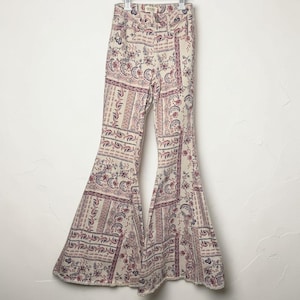 May include: Cream-colored, high-waisted bell-bottom pants with a floral and paisley pattern in shades of purple, red, and blue. The pants are hanging on a hanger against a white background. The fabric appears to be a lightweight material.