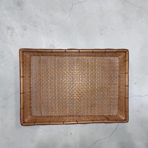 May include: A rectangular woven basket with a light brown color. The basket has a tightly woven pattern and a sturdy frame. The basket is empty and sits on a gray surface.
