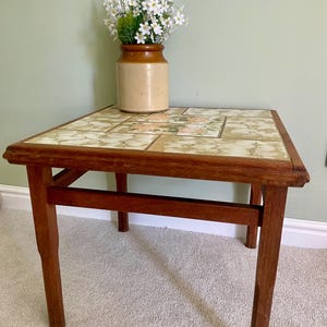 May include: A square wooden side table with a tiled top featuring a cream and brown marbled pattern and a central floral design. A two-toned brown ceramic jar filled with white artificial flowers sits on the table surface.