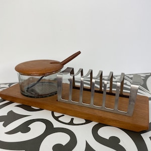 May include: A rectangular wooden serving tray featuring a clear glass condiment jar with a wooden lid and a matching wooden spoon, alongside a silver-toned metal toast rack with six vertical slots for holding slices of bread upright.