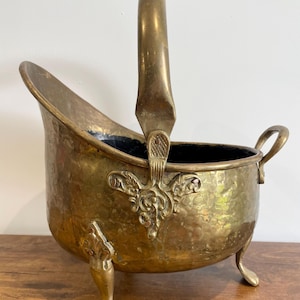 May include: A brass coal scuttle featuring a hammered texture, a tall arched handle, and three claw-style feet. The vessel has an open top, a decorative metal plate where the handle attaches to the body, and a small handle on the back side.