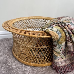 May include: A round, light brown wicker basket with a diamond-patterned lattice weave and a wide, flat rim. A folded, multi-colored woven throw blanket with a detailed floral and botanical pattern and fringed edges rests inside the basket.