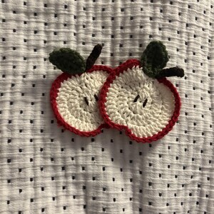 May include: Two crocheted apple slices with red edges, white centres, green leaves, and brown stems. The handmade coasters are placed on a white fabric with a black polka dot pattern. The apple slices are a charming, decorative item.