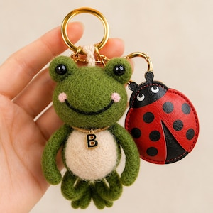 Può includere: Un portachiavi a forma di rana in feltro verde fatto a mano con un ciondolo iniziale color oro e un ciondolo coccinella rosso e nero. La rana ha guance rosa e occhi neri. Il portachiavi ha un anello color oro.
