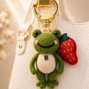 May include: A green frog keyring with a white belly, pink cheeks, and black eyes. The frog is holding a red strawberry. The keyring has a gold-coloured clasp and a small gold-coloured charm with the letter "B".
