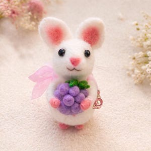 May include: A handcrafted white felt mouse figure with pink detailing on the ears, nose, and feet. The mouse holds a bunch of purple grapes and has a pink ribbon. A small charm is attached to the mouse.