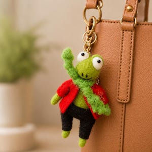 May include: A handmade frog keychain made of felt. The frog is green with large white eyes, wearing a red jacket, green scarf, and black pants. The keychain is attached to a gold-colored metal ring, hanging next to a brown leather bag.