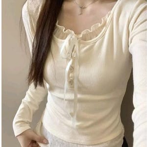 May include: Cream-coloured long-sleeve top with a ribbed texture. The top features a scoop neckline with a ruffled trim and a tie closure with two buttons. The bottom layer has a different, textured fabric.