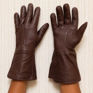 May include: A pair of dark brown leather gloves featuring a smooth finish, five fingers, and an extended cuff design that reaches past the wrist. The gloves are constructed with visible stitching along the fingers and a clean, minimalist aesthetic.