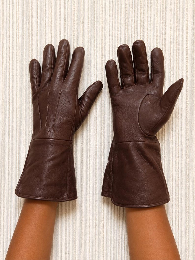 May include: A pair of dark brown leather gloves featuring a smooth finish, five fingers, and an extended cuff design that reaches past the wrist. The gloves are constructed with visible stitching along the fingers and a clean, minimalist aesthetic.