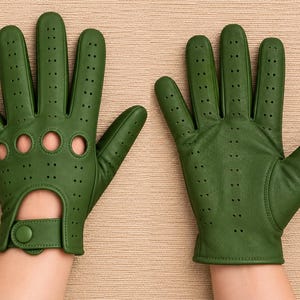 May include: A pair of olive green driving gloves featuring perforated detailing across the fingers and knuckles, with circular cutouts on the knuckles and a wrist strap secured by a snap button closure on one of the gloves.