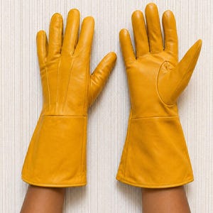 May include: A pair of mustard yellow leather gloves featuring long, flared cuffs that extend past the wrist. The gloves have visible stitching details along the back of the hand and fingers, with a smooth, matte finish throughout the material.