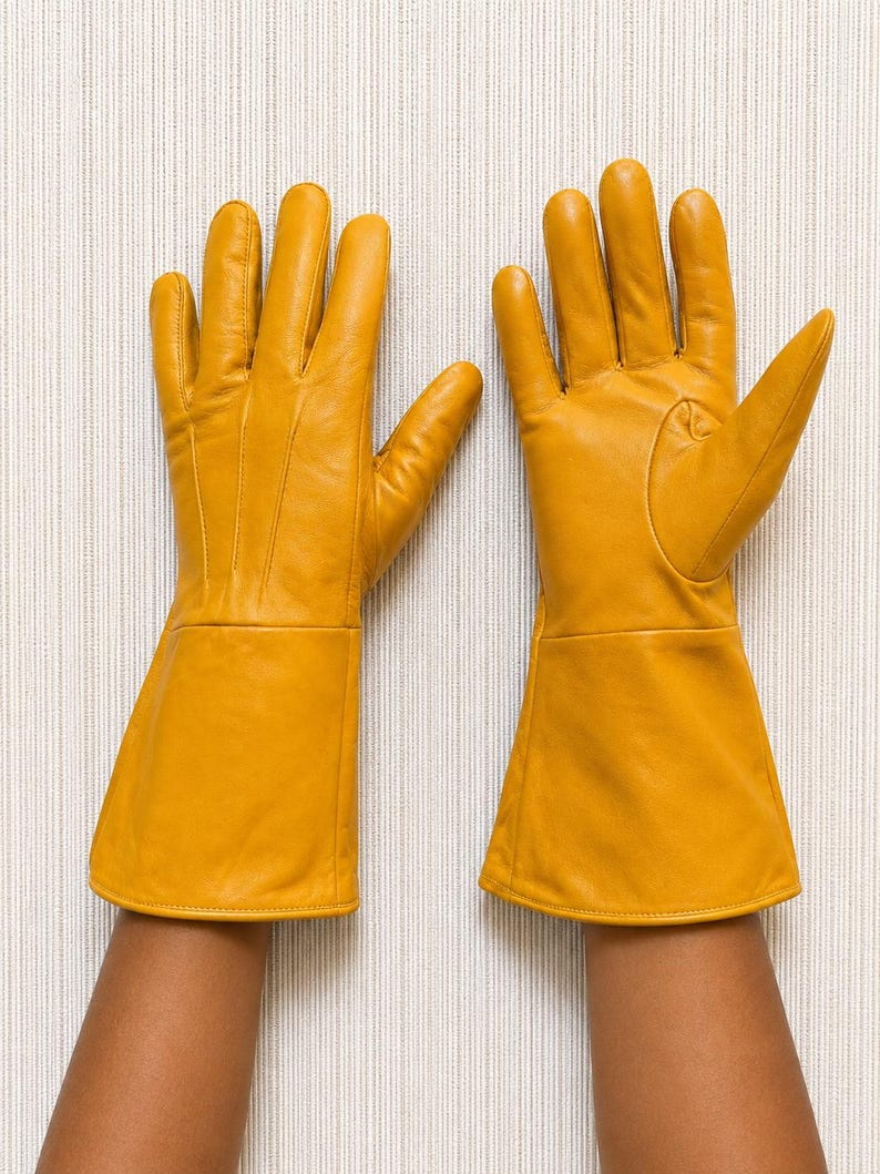 May include: A pair of mustard yellow leather gloves featuring long, flared cuffs that extend past the wrist. The gloves have visible stitching details along the back of the hand and fingers, with a smooth, matte finish throughout the material.