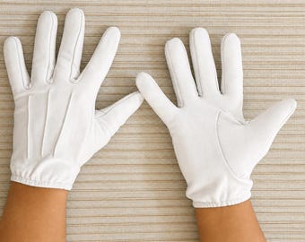 May include: A pair of white gloves featuring three stitched lines on the back of the hand, a gathered elastic wrist cuff, and a smooth finish. The gloves are designed with individual finger stalls and a thumb section, providing full hand coverage.