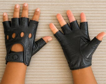 May include: A pair of black fingerless leather gloves featuring circular cutouts across the knuckles, perforated detailing on the fingers, and a wrist strap with a snap button closure. The gloves expose the fingers and thumb for dexterity.